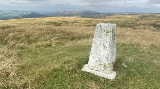 Open moorland on Merryton Low with a trig pillar on the summit and the distant ridge of The Roaches beyond